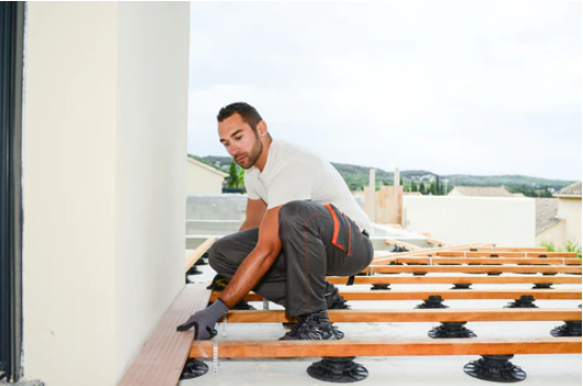 Ein Handwerker verlegt eine Bambus-Terrassendiele auf einer Unterkonstruktion, die mit höhenverstellbaren Stelzlagern montiert ist. Die Arbeiten finden an der Fassade eines Hauses statt.