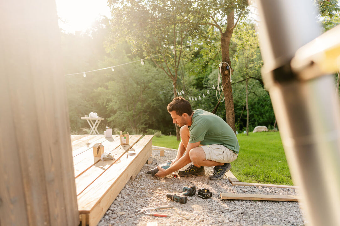 Montage einer Bambus-Terrasse im Garten: Ein Heimwerker befestigt die Unterkonstruktion für Bambus-Terrassendielen auf einem stabilen Splittbett. Die natürliche Umgebung und hochwertige Materialien unterstreichen die Nachhaltigkeit und Langlebigkeit von Bambus als Terrassenbelag.