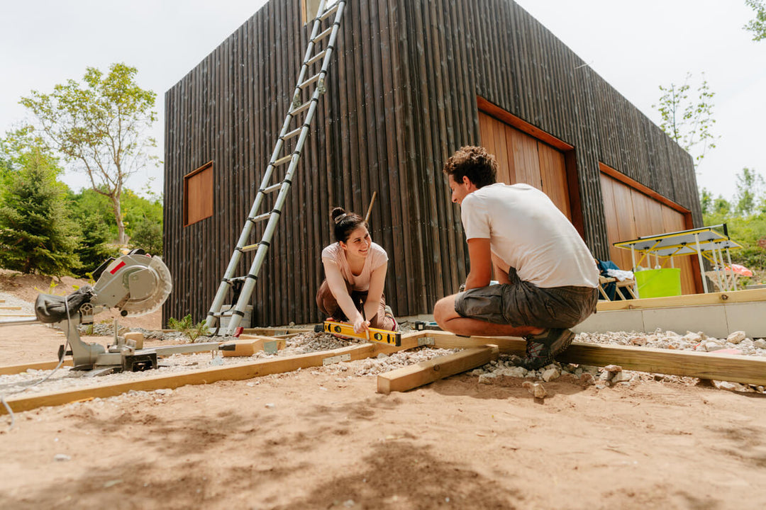 Ein Paar bereitet gemeinsam den Unterbau für ein aMbooo Terrassen Komplettpaket vor. Auf einem verdichteten Splittbett werden Holzträger mit einer Wasserwaage ausgerichtet – die Basis für die spätere Verlegung von langlebigen Bambus-Terrassendielen.
