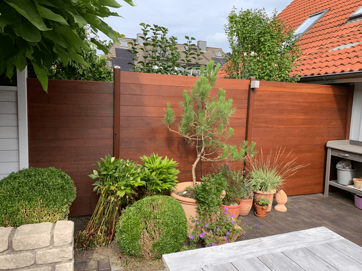 Gartenszene mit einem hohen, rotbraunen Bambus Sichtschutzzaun aus horizontalen Holzlatten. Davor sind diverse Pflanzen arrangiert, darunter eine kleine Kiefer in einem großen Terrakotta-Topf, runde Buchsbäume, Ziergräser und Stauden. Am rechten Bildrand steht ein grauer Pflanztisch aus Holz; im Hintergrund sind Hausdächer und Baumwipfel zu sehen.