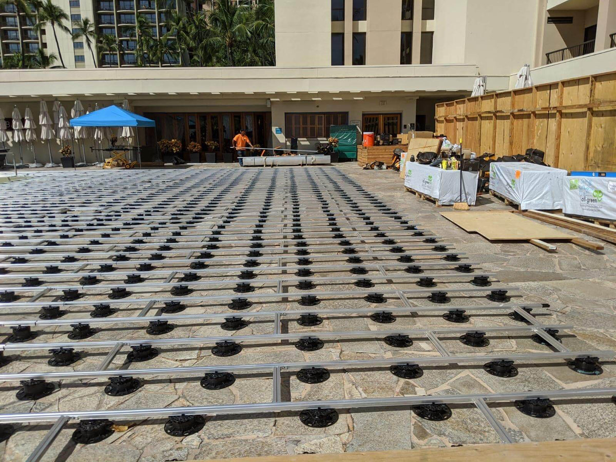 Terrassen-Unterkonstruktion aus Aluminium auf Stelzlagern vor einer roten Klinkerwand. Die Profile sind auf Betonplatten ausgerichtet und für die Verlegung von Terrassendielen vorbereitet.