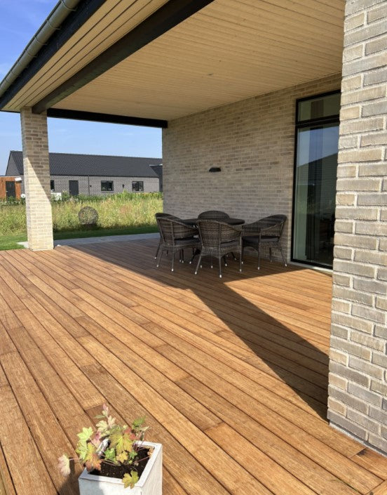 Überdachte Terrasse eines modernen Einfamilienhauses mit warmen Bambus Terrassendielen. Unter dem Dach steht eine Sitzgruppe aus vier Korbstühlen und einem runden Tisch. Die Terrasse grenzt an eine helle Klinkerfassade mit großen Fensterflächen und bietet Blick auf ein begrüntes Grundstück sowie benachbarte Neubauten.