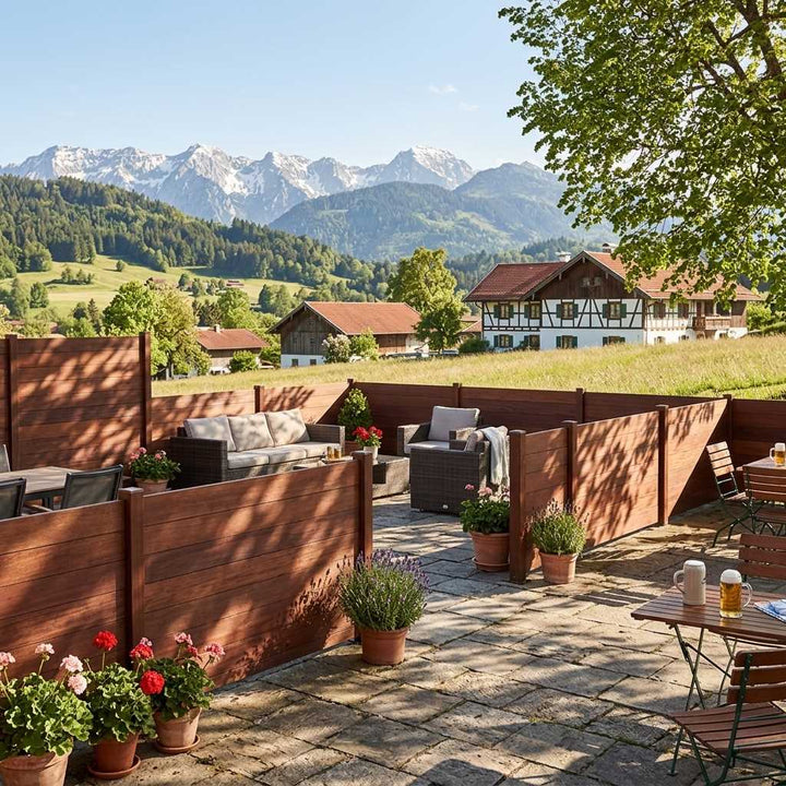 Terrasse mit coffee farbenden Holz-Sichtschutzwänden, Sitzmöbeln und Topfpflanzen; dahinter liegen Wiesen, Bauernhäuser und ein Alpenpanorama.