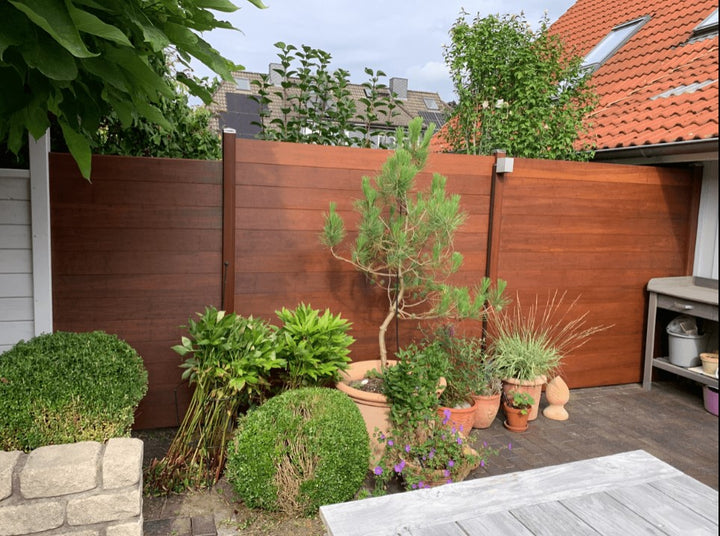 ModernerAMBOOO Bambus-Sichtschutzzaun in warmer Optik mit dunklen Pfosten, der eine Terrasse begrenzt und als Hintergrund für diverse Gartenpflanzen und einen Pflanztisch dient.