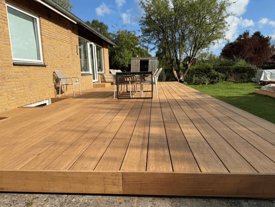 Große Bambusholz Terrasse in der Farbe Sand mit modernen Gartenmöbeln vor einem Einfamilienhaus, umgeben von Rasen und Bäumen bei sonnigem Wetter.