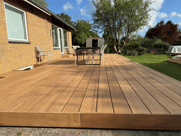Große Bambusholz Terrasse in der Farbe Sand mit modernen Gartenmöbeln vor einem Einfamilienhaus, umgeben von Rasen und Bäumen bei sonnigem Wetter.