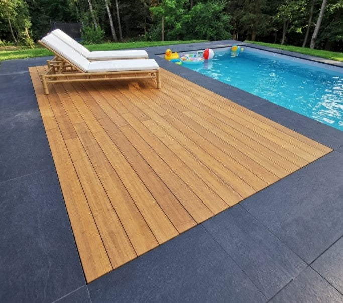 Pool mit warmer Holzterrasse, dunklen Steinplatten und zwei weißen Sonnenliegen vor einem grünen Wald.
