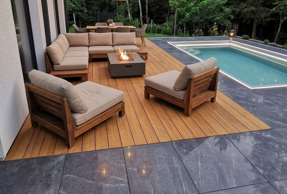 Pool mit warmer Holzterrasse, dunklen Steinplatten und zwei weißen Sonnenliegen vor einem grünen Wald.