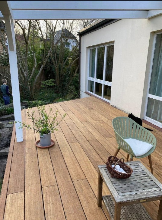 Moderne Terrasse mit Sand Bambus-Terrassendielen unter einer weißen Pergola. Die Dielen sind parallel zur Hauswand verlegt und erzeugen eine natürliche, warme Holzoptik. Auf der Terrasse stehen ein kleiner Holztisch mit Dekoration, ein grüner Stuhl mit Sitzkissen sowie eine Topfpflanze. Große Fensterfronten und ein Garten mit Bäumen bilden den Hintergrund.