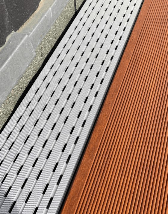 Silberfarbene Terrassen-Drainage mit länglichen Entwässerungsschlitzen neben braunen geriffelten Holzdielen.