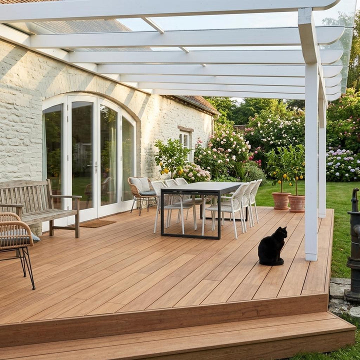 Überdachte Closeddecking Holzterrasse mit Esstisch, Sitzmöbeln und schwarzer Katze vor einem hellen Landhaus mit blühendem Garten.