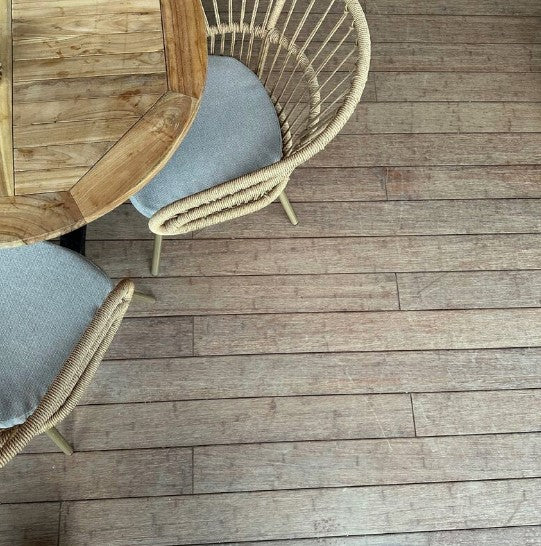 Terrassenboden Cmapoverde in vergrauter Sandoptik mit längs verlegten Dielen. Im oberen linken Bildbereich stehen ein Holztisch und zwei Outdoor-Stühle aus Rattan mit hellgrauen Sitzkissen. 
