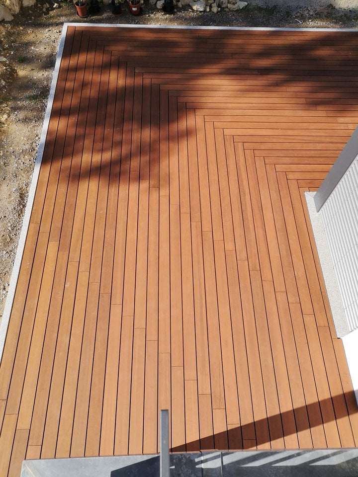 Große Terrasse aus hellbraunen Holzdielen, die in einem dekorativen Fischgrätmuster um die Ecke eines Gebäudes verlegt wurden. Die schmalen Dielen sind von einem hellen Rand eingefasst. Auf der Terrasse fallen Schatten von Bäumen, wodurch die hochwertige Verlegearbeit besonders gut sichtbar wird.