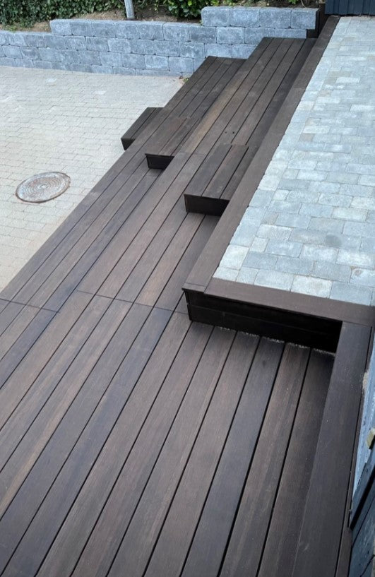 Die Aufnahme zeigt eine moderne Terrassenanlage mit dunkelbraunen Terrassendielen in geradliniger Verlegung. In die erhöhte Terrasse sind mehrere Sitzstufen und Sitzbänke integriert, die sich harmonisch in das Design einfügen. Angrenzend befinden sich helle Pflasterflächen und eine Natursteinmauer, die den Außenbereich strukturiert und der Anlage ein hochwertiges, modernes Erscheinungsbild verleiht.