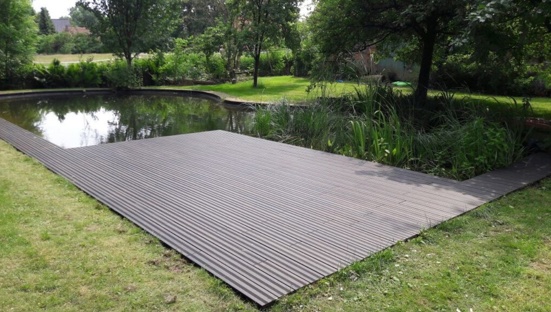 Eine weitläufige Terrasse mit schwarzen, fein geriffelten Terrassendielen erstreckt sich entlang eines naturnah gestalteten Gartenteichs. Das ruhige Wasser spiegelt die umliegenden Bäume und Sträucher wider. Rund um den Teich sorgen Rasenflächen, Schilfpflanzen und dichtes Grün für eine harmonische Gartenatmosphäre. Die Terrasse bietet einen direkten Zugang zum Wasser und fügt sich nahtlos in die natürliche Umgebung ein.
