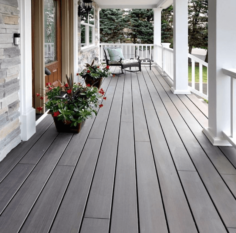 Eine überdachte Veranda mit schwarzen Ambooo Terrassendielen erstreckt sich entlang der Hausfassade. Weiße Säulen und Geländer rahmen den Außenbereich ein. Neben der Eingangstür stehen bepflanzte Blumenkübel, während am Ende der Veranda eine Sitzecke mit Sesseln zum Entspannen einlädt. Im Hintergrund sind Bäume und Gartenflächen zu sehen.