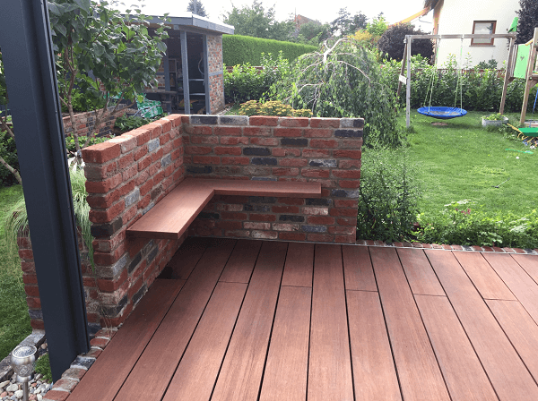 Moderne Terrasse mit rötlich-braunen Bambus-Terrassendielen und einer integrierten Sitzecke aus Klinkermauerwerk im gepflegten Garten.